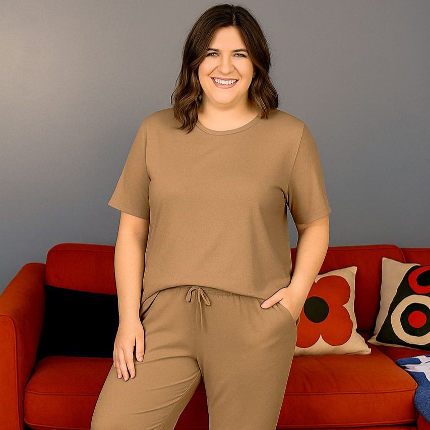 A woman wearing a brown short sleeve breastfeeding loungewear set, standing in front of a red couch with colorful cushions.