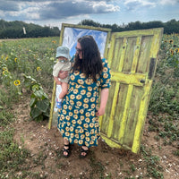 Woman stood on a grassy field with a wooden box, surrounded by sunflowers and blue sky.