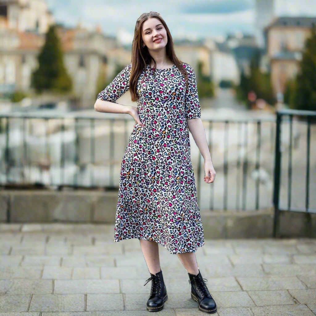Stylish Mum with hand on hip with rainbow leopard print breastfeeding dress. She has long dark here and is posing confidently. She is also black boots and is stood in on a pavement on a street.