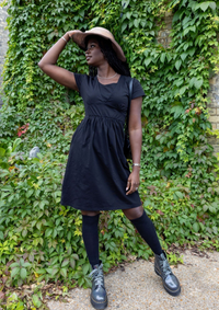 Woman in a black breastfeeding dress and hat standing in front of a green ivy-covered wall.