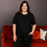 A woman wearing a black short sleeve breastfeeding loungewear set, standing in front of a red couch with a white and orange cushion.
