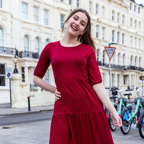 Stylish Mum with dark hair pinned up. She has her hand on her hip and has a huge smile. She is wearing a stunning red midi dress which has zips for breastfeeding. She is wearing black tights and boots. The dress is made by Stylish Mum who makes breastfeeding dresses in the UK. 