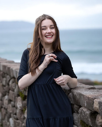 Woman in a black breastfeeding dress standing against a stone wall with ocean in the background showing breastfeeding access via a zip.
