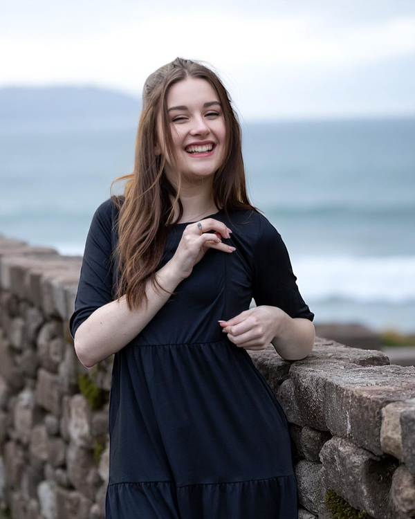 Woman in a black breastfeeding dress standing against a stone wall with ocean in the background showing breastfeeding access via a zip.