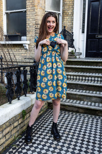 Woman in a floral breastfeeding dress standing on a checkered pavement outside a building. She is showing the breastfeeding access of the dress with her hands. 