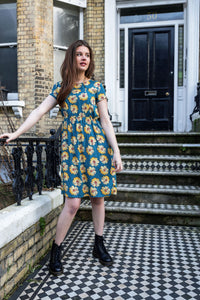 A model wearing a knee-length blue breastfeeding dress with a sunflower pattern, featuring short sleeves and a Peter Pan collar, standing on a doorstep.