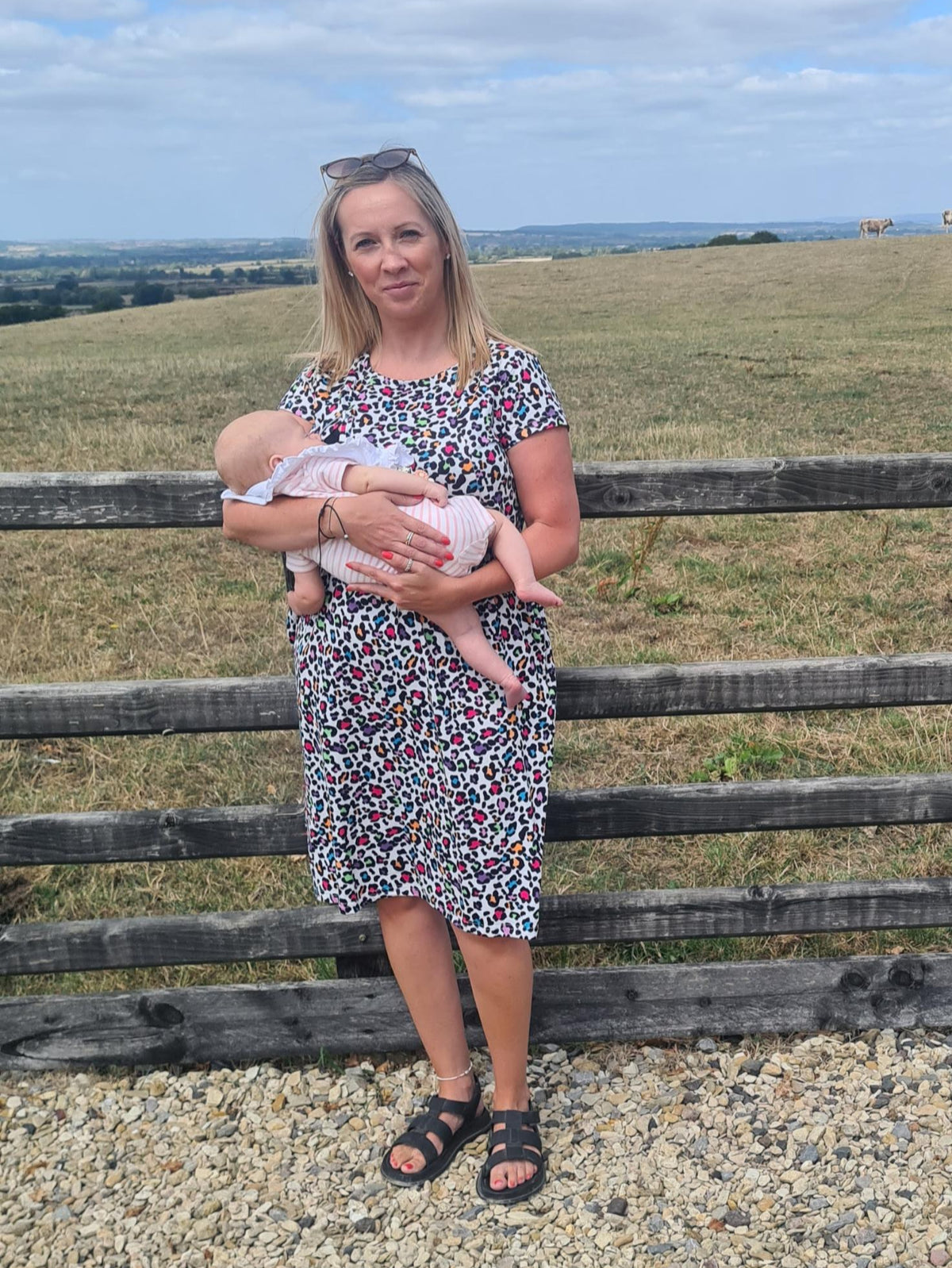 Woman breastfeeding a baby in a patterned breastfeeding dress standing near a wooden fence with a scenic background.