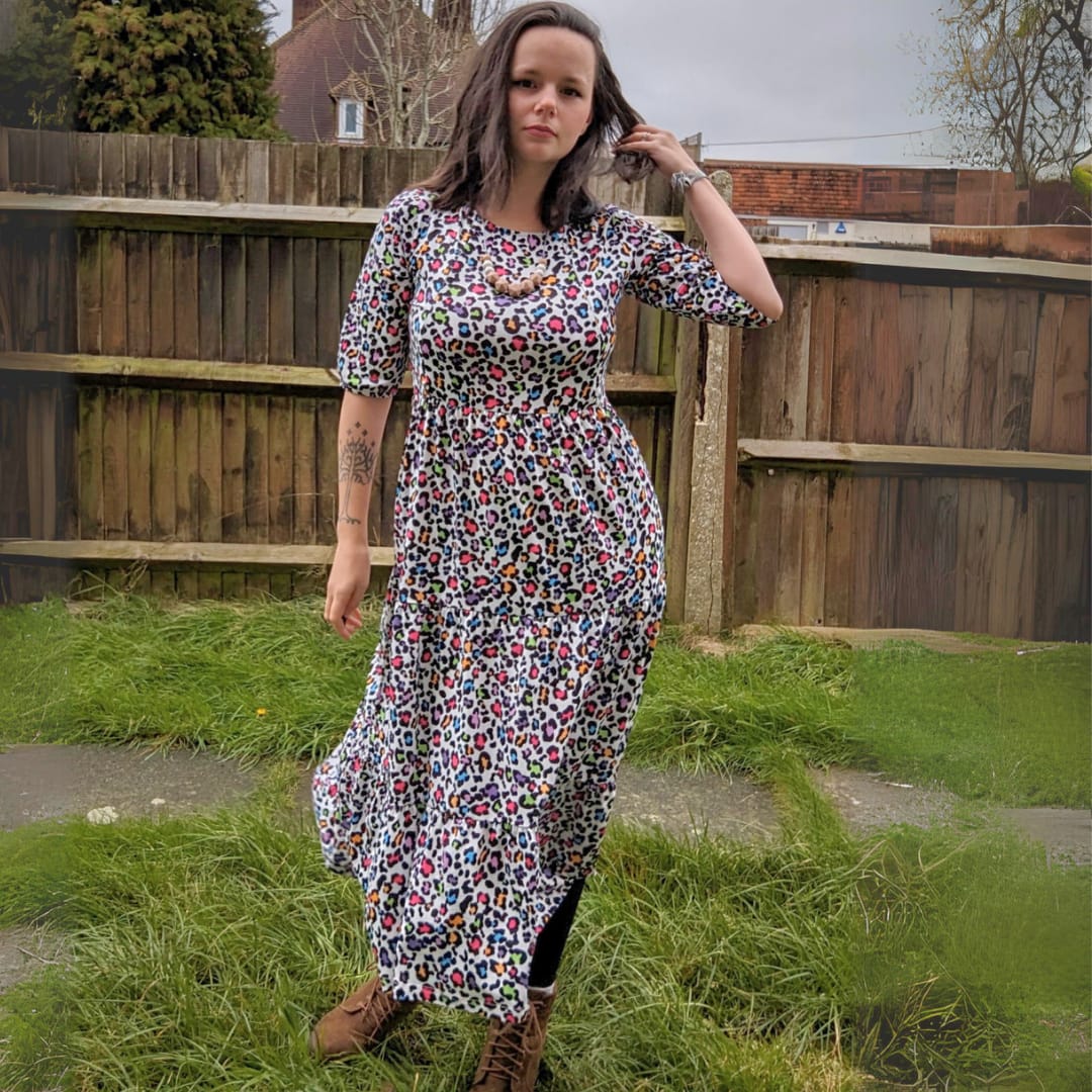 A mum in a garden in a rainbow leopard print nursing dress. The mum has one hand by her side and the other brushing her dark hair back. She is also wearing brown boots and a chunky teething necklace.